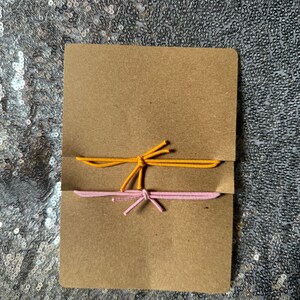 May include: Two elastic hair ties, one yellow and one pink, are tied together and displayed on a brown card.