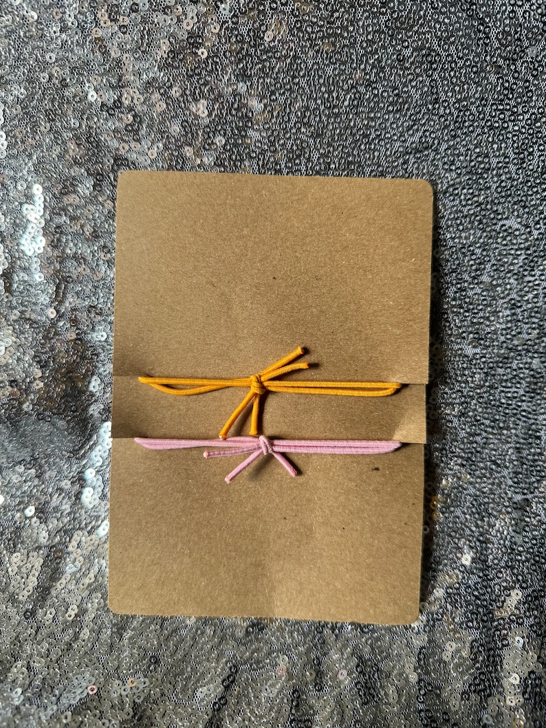 May include: Two elastic hair ties, one yellow and one pink, are tied together and displayed on a brown card.