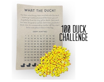 May include: A brown card with the text "What the Duck!" and "100 Duck Challenge" printed on it. A chart with numbers 1 through 100 is printed on the card. A pile of 100 yellow rubber duck toys is scattered on the card.