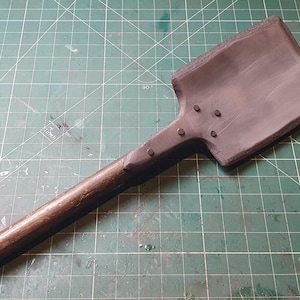 May include: A dark gray shovel with a wooden handle. The shovel head is rectangular and attached to the handle with rivets. The handle is a dark brown color. The shovel is lying on a green cutting mat.