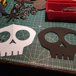 May include: Two skull-shaped stencils, one white and one black, are laid out on a green cutting mat. The white stencil has the number "150" written on it.