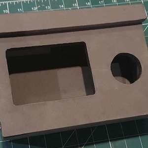 Pode incluir: Uma caixa de cartão castanho com duas aberturas recortadas. A abertura maior é retangular e a menor é redonda.