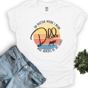 Könnte beinhalten: Weißes T-Shirt mit einem Grafikdruck eines Sonnenuntergangs und eines Elchs. Der Text "No matter where I roam, Dildo will always be my home!" ist auf dem T-Shirt aufgedruckt.
