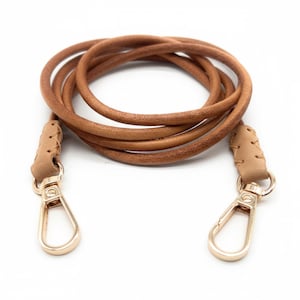 Könnte beinhalten: Eine aufgerollte, hellbraune Lederriemen mit goldfarbenen Metallverschlüssen. Der Riemen ist für eine Tasche oder einen Geldbeutel konzipiert, mit geflochtenen Lederakzenten an jedem Ende, an dem die Verschlüsse befestigt sind.