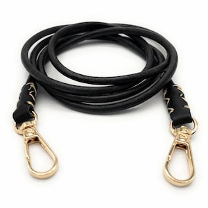 Può includere: Tracolla per borsa in pelle nera con chiusure in metallo color oro. Il cinturino è realizzato con un cordino rotondo in pelle intrecciata. Le estremità del cinturino sono attaccate alle chiusure con pelle nera e cuciture dorate.