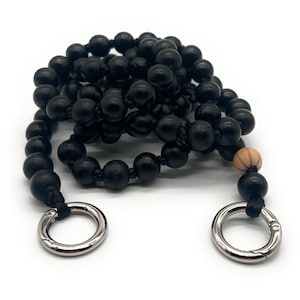 Könnte beinhalten: Schwarzer Perlenriemen mit silberfarbenen Metallringen. Der Riemen besteht aus kleinen, runden, schwarzen Perlen. Ein Ende hat eine hellbraune Holzperle. Der Riemen ist für die Befestigung an einer Tasche oder einem Beutel konzipiert.