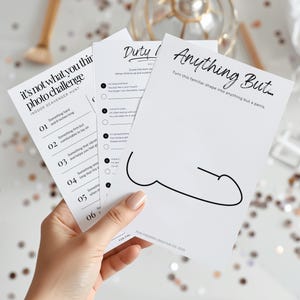 Op de afbeelding: Een set van drie witte papieren spelkaarten met zwarte tekst en afbeeldingen. De bovenste kaart bevat de tekst Anything But... en een lijntekening van een fallische vorm. Andere kaarten bevatten de tekst It's not what you think photo challenge en Dirty.