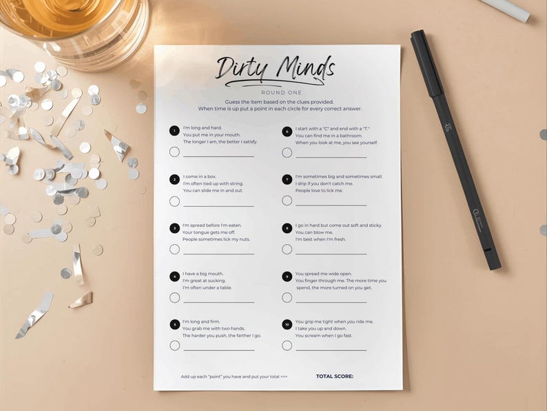 May include: A printed game sheet titled "Dirty Minds" with fill-in-the-blank clues, a black pen, and scattered confetti. The game sheet is on a light beige surface, with a glass of liquid visible in the upper left corner.