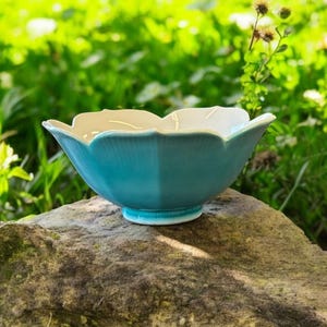 Könnte beinhalten: Eine türkisfarbene Keramikschale mit weißem Innenraum, geformt wie eine Blume, steht auf einem großen Felsen. Die Schale hat einen Sockel und ist von grünem Laub und Sonnenlicht umgeben.