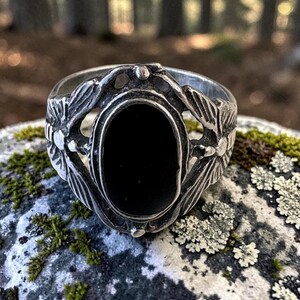 May include: A silver ring featuring a central oval black stone set within a decorative metal frame. The band displays intricate, textured wing-like motifs on both sides of the stone, creating a detailed, symmetrical design with a polished finish.