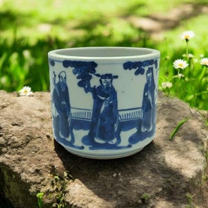 Puede incluir: Una taza de té de cerámica blanca con un diseño azul y blanco de cuatro figuras con vestimenta tradicional china. Las figuras están rodeadas de un diseño floral azul y blanco.