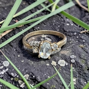 May include: A gold ring featuring a central oval-cut light blue gemstone flanked by three smaller round-cut clear stones on each side. The band is polished gold and includes a visible hallmark stamp on the interior surface of the metal band.