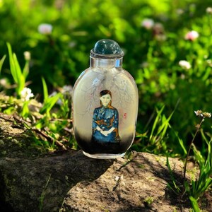 Può includere: Un'ampolla di vetro trasparente con un tappo verde. L'ampolla presenta un'immagine dipinta di una donna in un abito blu e oro. Lo sfondo è un'immagine sfocata di un albero e dei rami. L'ampolla è appoggiata su un tronco.