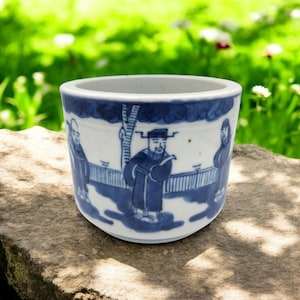Puede incluir: Una taza de cerámica blanca con un diseño azul y blanco de tres figuras con ropa tradicional china. Las figuras están de pie frente a un árbol y una valla.