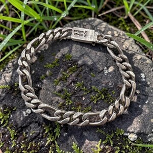 Puede incluir: Una pulsera de cadena de metal plateado con eslabones ovalados y circulares entrelazados. La pulsera incluye un cierre de caja rectangular con el texto grabado 925 y caracteres adicionales visibles en la superficie del cierre metálico.