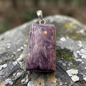 May include: A rectangular pendant featuring a polished purple and white swirled stone with dark inclusions, set in a dark metal frame with a silver-toned bail at the top for attachment to a necklace chain.
