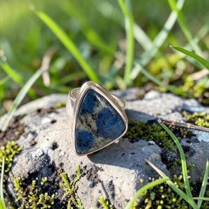 Op de afbeelding: Een ring met een driehoekige steen met een glad, gepolijst oppervlak. De steen vertoont een gevlekt patroon van diepblauwe en grijs-witte kleuren. De steen is gezet in een eenvoudige, gepolijste zilverkleurige metalen band en zetting.