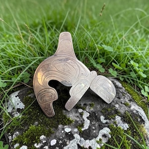May include: A metallic brooch shaped like an orca, featuring intricate engraved line work and patterns across its surface. The design depicts the stylised form of a whale with a prominent dorsal fin and detailed markings throughout the body.