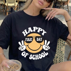 May include: A black t-shirt featuring a graphic of a yellow smiley face wearing black sunglasses, making peace signs with both hands, and surrounded by the white text "HAPPY FIRST DAY OF SCHOOL" in a bold, rounded, retro-style font.