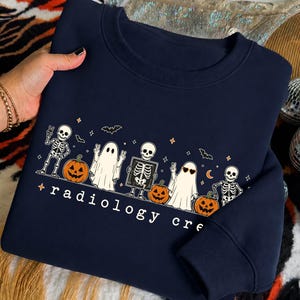 Peut inclure: Un sweat-shirt bleu marine à col rond avec une impression graphique de squelettes, de fantômes et de citrouilles d'Halloween. Le motif inclut le texte "radiology crew" en lettres minuscules blanches, avec des étoiles, des chauves-souris et une lune.
