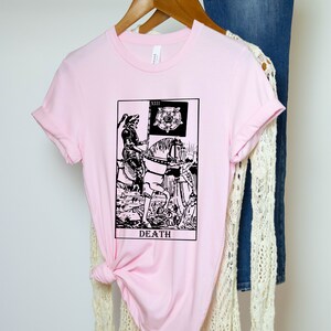 Pode incluir: Camiseta rosa com uma ilustra&ccedil;&atilde;o em preto e branco do cart&atilde;o de tar&ocirc; da Morte. O cart&atilde;o mostra uma figura esquel&eacute;tica montando um cavalo e segurando uma bandeira preta com uma rosa branca.