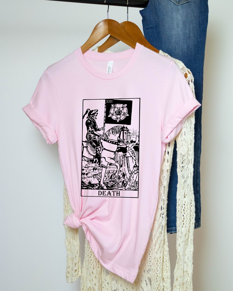 Pode incluir: Camiseta rosa com uma ilustra&ccedil;&atilde;o em preto e branco do cart&atilde;o de tar&ocirc; da Morte. O cart&atilde;o mostra uma figura esquel&eacute;tica montando um cavalo e segurando uma bandeira preta com uma rosa branca.