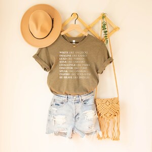 May include: A green t-shirt with white text that reads "Write like Angelou, Imagine like Kahlo, Lead like Tubman, Soar like Earhart, Challenge like Parks, Discover like Curie, Speak like Ginsburg, Inspire like Yousafzai, Be Brave like Bridges".
