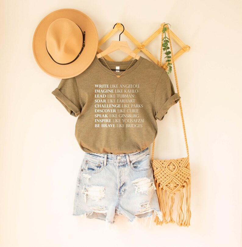 May include: A green t-shirt with white text that reads "Write like Angelou, Imagine like Kahlo, Lead like Tubman, Soar like Earhart, Challenge like Parks, Discover like Curie, Speak like Ginsburg, Inspire like Yousafzai, Be Brave like Bridges".