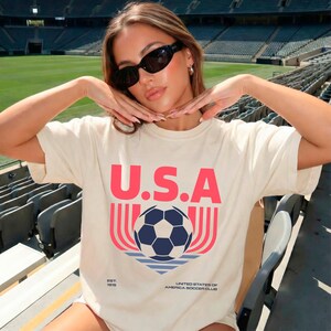 May include: Cream-colored t-shirt with the word "U.S.A." in red block letters above a soccer ball graphic. The graphic includes a blue and white soccer ball, red and pink lines, and the text "UNITED STATES OF AMERICA SOCCER CLUB".