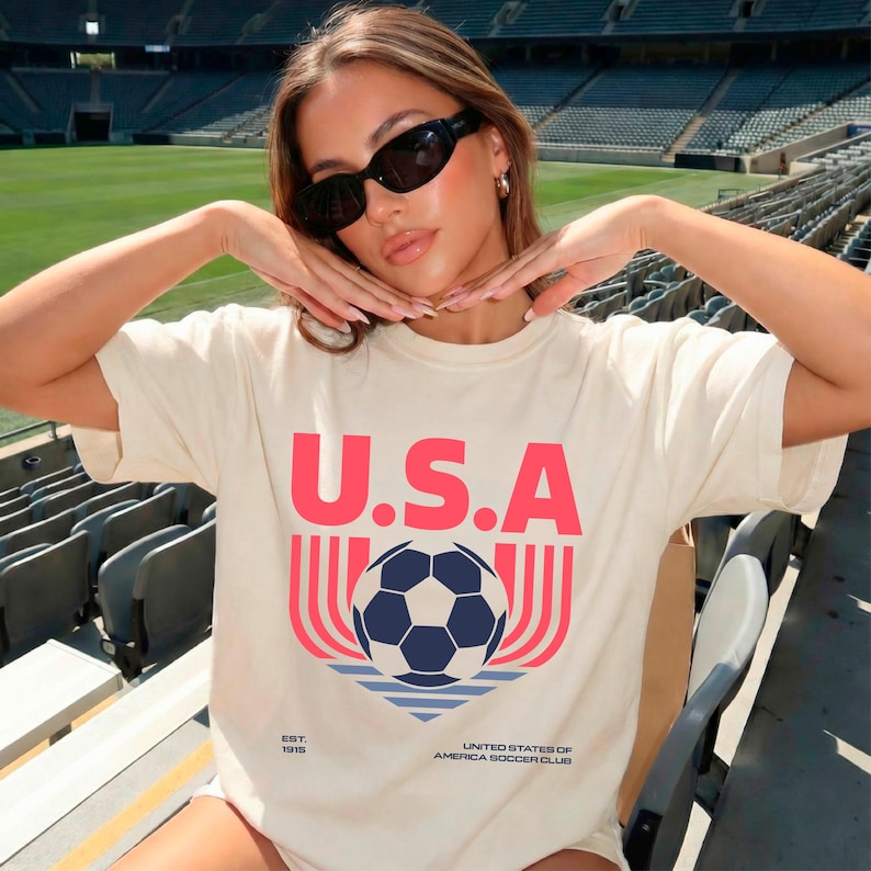 May include: Cream-colored t-shirt with the word "U.S.A." in red block letters above a soccer ball graphic. The graphic includes a blue and white soccer ball, red and pink lines, and the text "UNITED STATES OF AMERICA SOCCER CLUB".