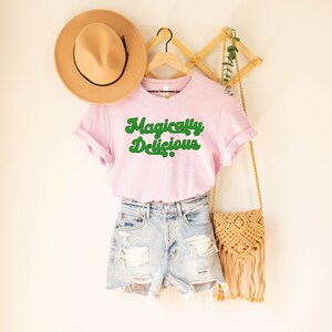 May include: A pink t-shirt with the text "Magically Delicious" in green lettering. The shirt is hanging on a wooden rack with a brown hat and a straw bag.