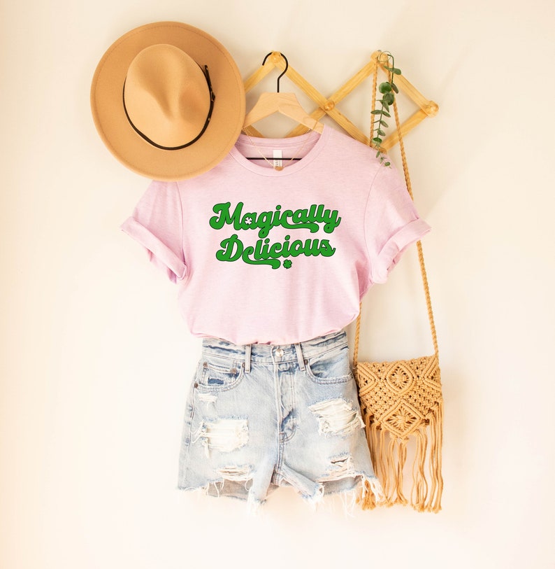 May include: A pink t-shirt with the text "Magically Delicious" in green lettering. The shirt is hanging on a wooden rack with a brown hat and a straw bag.