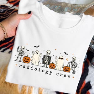 May include: A white crewneck t-shirt featuring a Halloween-themed graphic with skeletons, ghosts, and jack-o'-lanterns. The design includes the text "radiology crew" in black lowercase letters, accented by small orange stars and black bats.