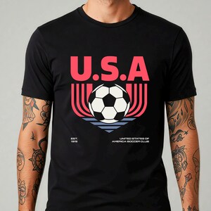 May include: Black t-shirt with a graphic design featuring the letters "U.S.A" in red above a soccer ball. Below the ball are red and blue lines, with the text "United States of America Soccer Club" and "Est. 1915".