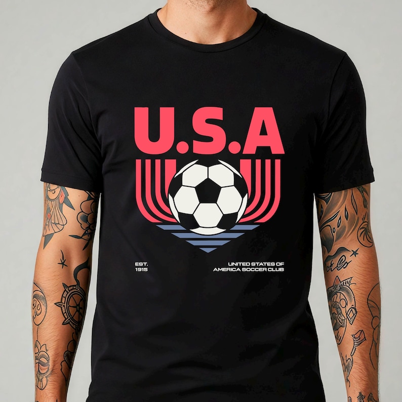 May include: Black t-shirt with a graphic design featuring the letters "U.S.A" in red above a soccer ball. Below the ball are red and blue lines, with the text "United States of America Soccer Club" and "Est. 1915".