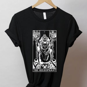 Pode incluir: Camiseta preta com um gr&aacute;fico branco do cart&atilde;o de tar&ocirc; O Hierofante. O cart&atilde;o apresenta uma figura sentada em um trono com uma chave e um cajado. O texto "THE HIEROPHANT" est&aacute; impresso abaixo do cart&atilde;o.