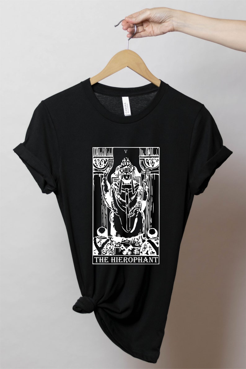 Pode incluir: Camiseta preta com um gr&aacute;fico branco do cart&atilde;o de tar&ocirc; O Hierofante. O cart&atilde;o apresenta uma figura sentada em um trono com uma chave e um cajado. O texto "THE HIEROPHANT" est&aacute; impresso abaixo do cart&atilde;o.