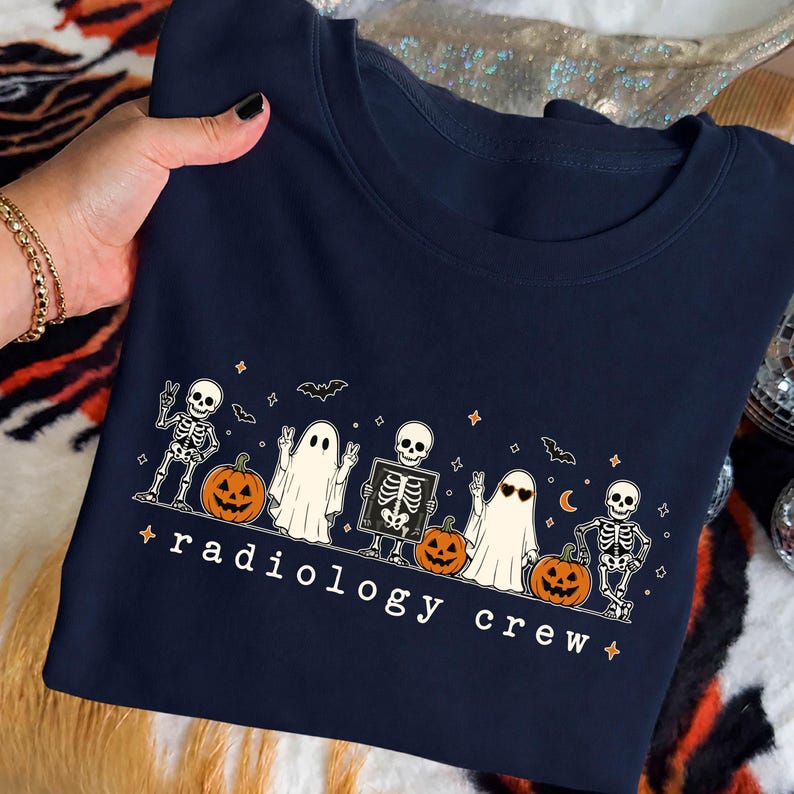 May include: A navy blue t-shirt featuring a graphic print of two skeletons, two ghosts, and three jack-o'-lanterns. One skeleton holds an X-ray image. The text "radiology crew" is printed in white below the characters, accented by small stars.