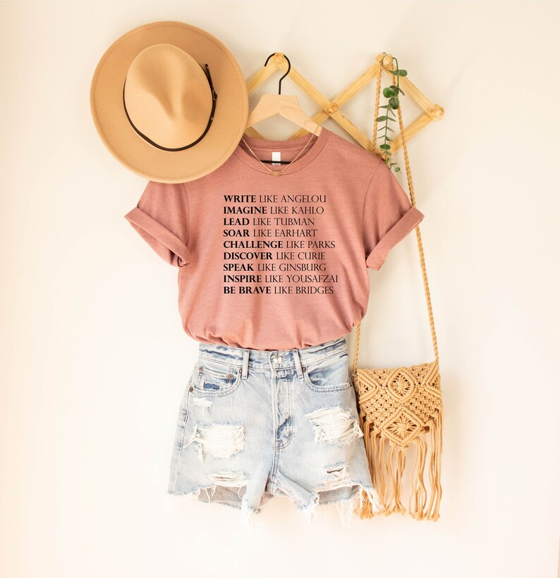 May include: A pink t-shirt with a black graphic print that reads "Write like Angelou, Imagine like Kahlo, Lead like Tubman, Soar like Earhart, Challenge like Parks, Discover like Curie, Speak like Ginsburg, Inspire like Yousafzai, Be Brave like Bridges".