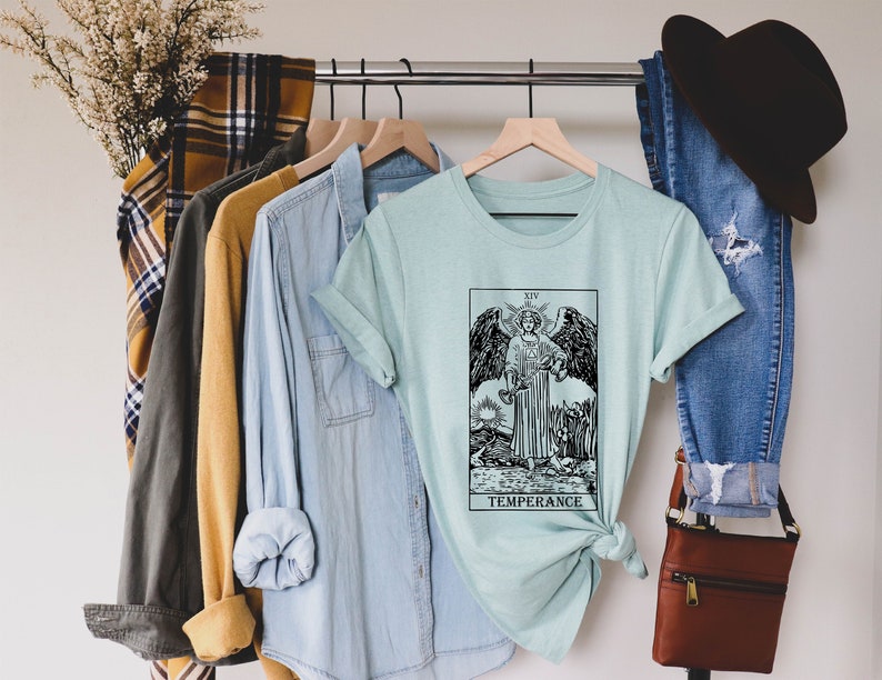 Pode incluir: Uma camiseta azul claro com uma ilustra&ccedil;&atilde;o em preto e branco do cart&atilde;o da Temperan&ccedil;a do baralho de tar&ocirc;. O cart&atilde;o mostra um anjo despejando &aacute;gua de uma ta&ccedil;a para outra. O texto "XIV TEMPERANCE" est&aacute; impresso abaixo da imagem.