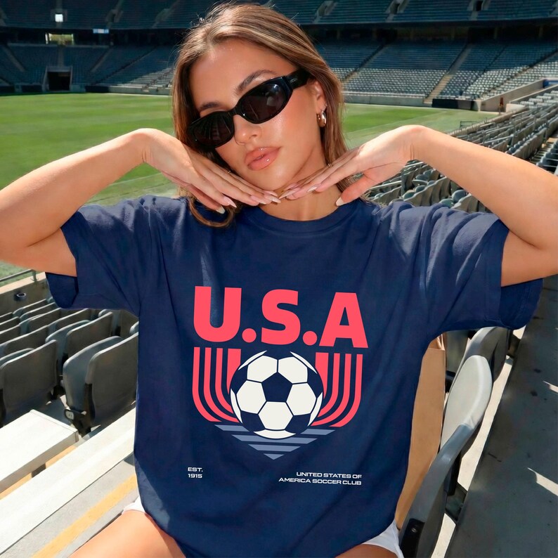 May include: Navy blue t-shirt with the text "U.S.A" in red block letters above a soccer ball graphic. The shirt also features the text "EST. 1919" and "UNITED STATES OF AMERICA SOCCER CLUB" in white.