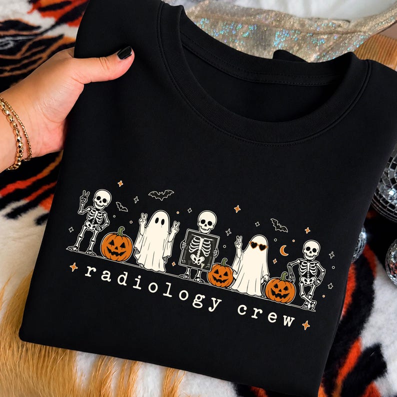May include: A black crewneck sweatshirt featuring a graphic print of skeletons, ghosts, and jack-o'-lanterns. The design includes the text "radiology crew" in a white font, accompanied by small star and bat illustrations across the chest area.