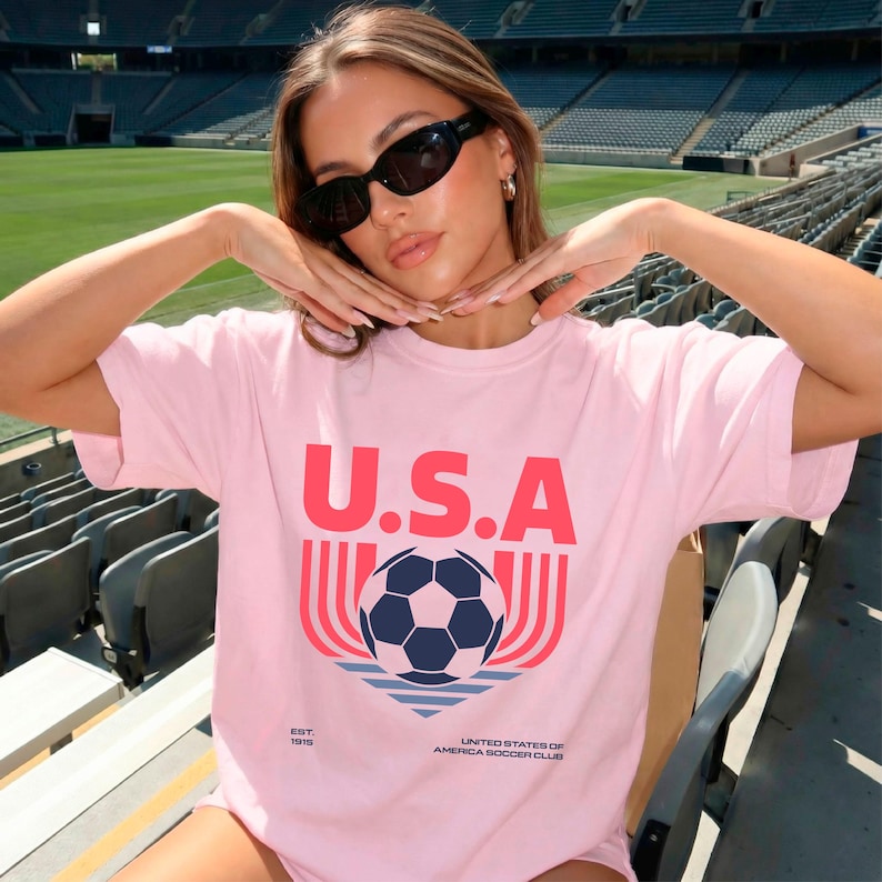 May include: A light pink t-shirt with the word "U.S.A" in red block letters above a soccer ball graphic. The graphic includes a blue and white soccer ball, red lines, and the text "UNITED STATES OF AMERICA SOCCER CLUB".