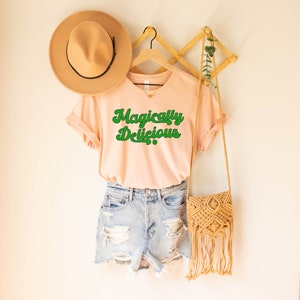 May include: A light pink t-shirt with the text "Magically Delicious" in green retro font. The shirt is hanging on a wooden hanger with a straw hat and a woven bag.