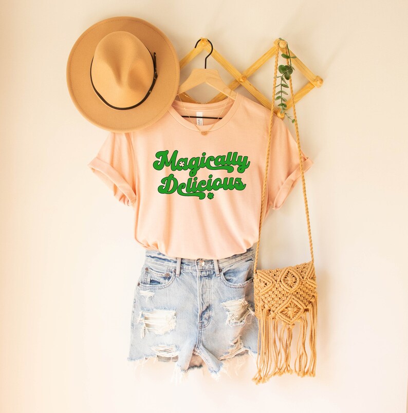 May include: A light pink t-shirt with the text "Magically Delicious" in green retro font. The shirt is hanging on a wooden hanger with a straw hat and a woven bag.