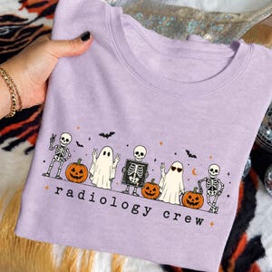 May include: A light purple t-shirt featuring a graphic print of skeletons, ghosts, and jack-o'-lanterns. The design includes the text "radiology crew" in black lowercase letters, along with small bats and orange star accents across the front.