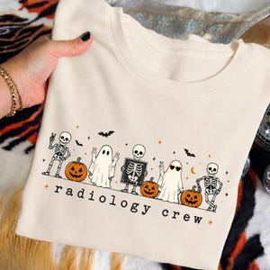 May include: A cream-colored t-shirt featuring a graphic print of skeletons, ghosts, and pumpkins. The design includes the text "radiology crew" in black lowercase letters, accompanied by small orange stars, bats, and a skeleton holding an X-ray.