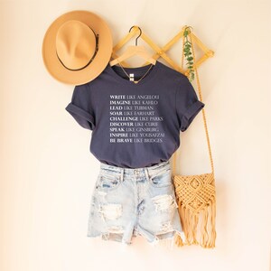 May include: A navy blue t-shirt with white text that reads "Write like Angelou, Imagine like Kahlo, Lead like Tubman, Soar like Earhart, Challenge like Parks, Discover like Curie, Speak like Ginsburg, Inspire like Yousafzai, Be Brave like Bridges".