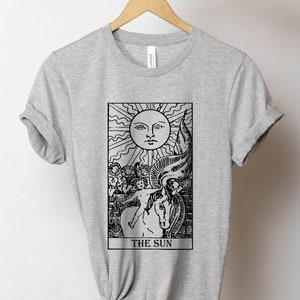 Pode incluir: Camiseta cinza com uma ilustra&ccedil;&atilde;o em preto e branco do cart&atilde;o de tar&ocirc; O Sol. O cart&atilde;o mostra um sol com um rosto, uma crian&ccedil;a montando um cavalo branco e o numeral romano XIX.