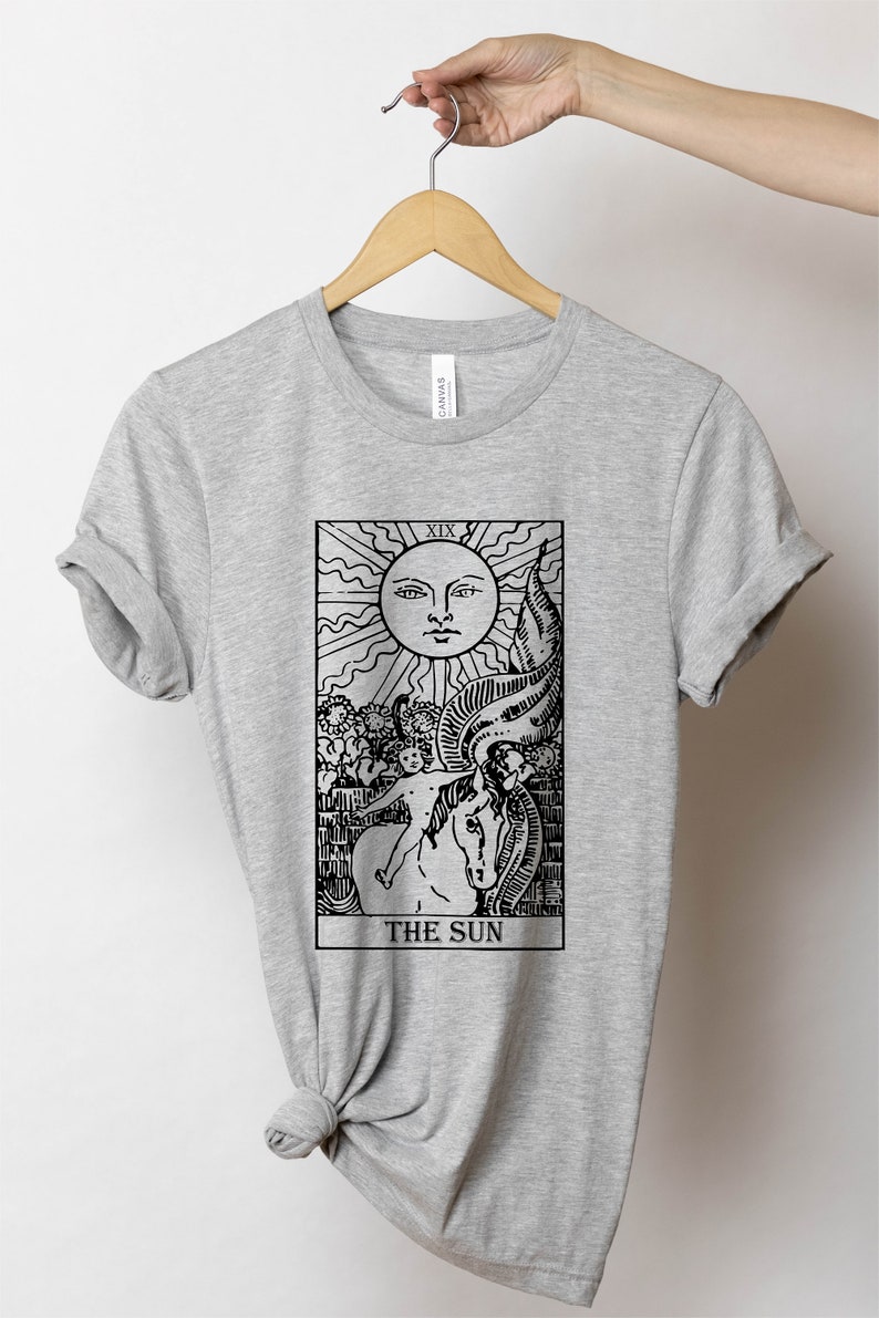 Pode incluir: Camiseta cinza com uma ilustra&ccedil;&atilde;o em preto e branco do cart&atilde;o de tar&ocirc; O Sol. O cart&atilde;o mostra um sol com um rosto, uma crian&ccedil;a montando um cavalo branco e o numeral romano XIX.